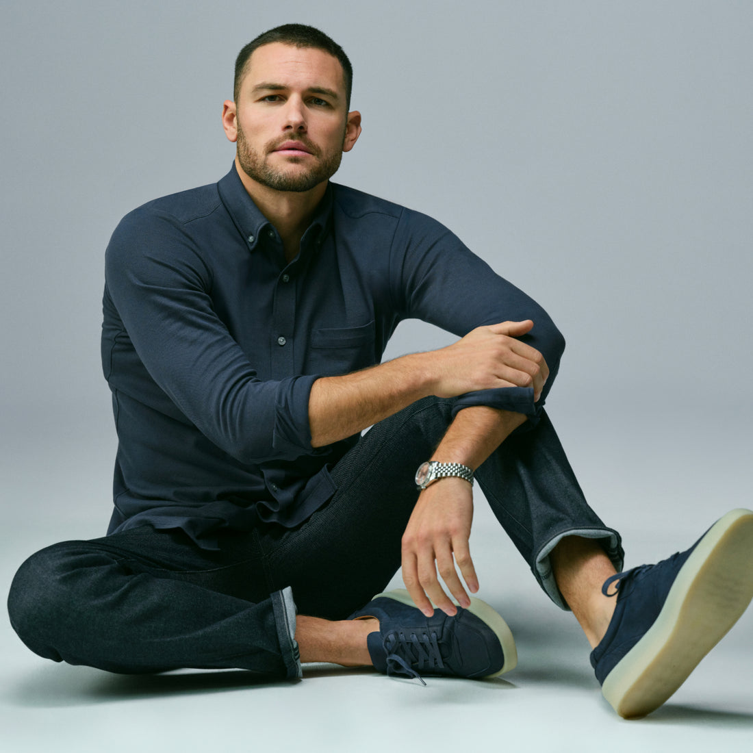 Man sits on the ground wearing a navy blue long sleeve button up and SILVER denim jeans