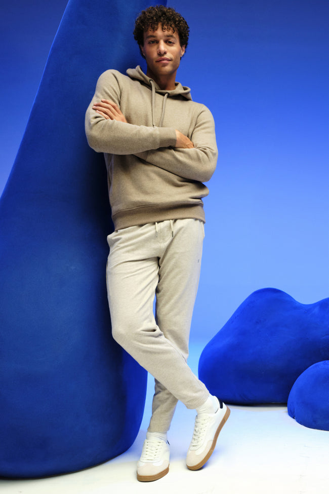 Man standing up against a blue backdrop with a cream hoodie and sweatpants.