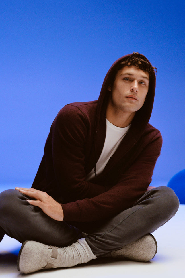 A man sits criss cross on the ground wearing a maroon zip up hoodie and sweatpants.