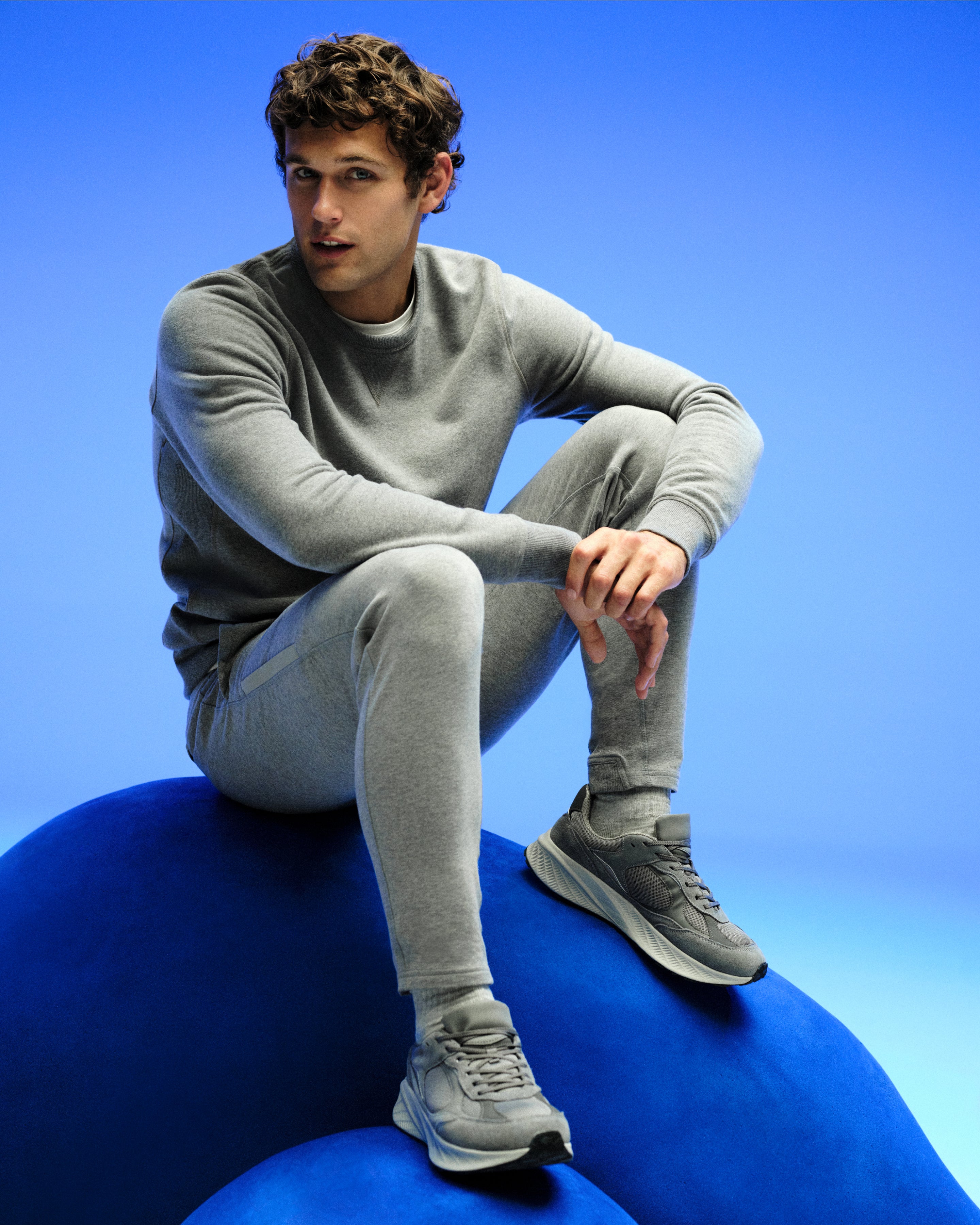 Man sits on a blue blob. he is wearing a grey sweatshirt and grey sweatpants